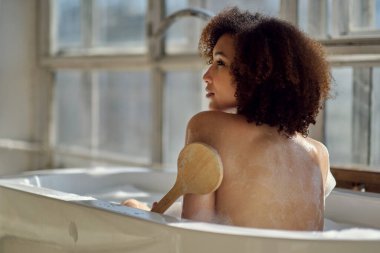 Gülümseyen ve rahatlatan Afro-Amerikan kadın köpük dolu bir küvette banyo yapıyor. Harika bir zaman. Yaşam tarzı insan kavramı