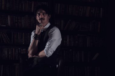 Karanlık bir kütüphane odasında düşünceli bir adam gece ayakta duruyor. Bıyıklı şık bir adam bileziği düzeltiyor..