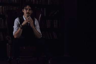 Karanlık bir kütüphane odasında düşünceli bir adam gece ayakta duruyor. Bıyıklı şık bir adam bileziği düzeltiyor..