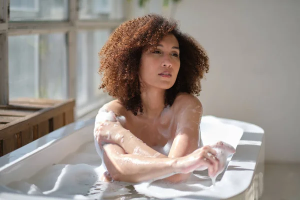 Krullend zwart meisje liggen, ontspannen in het bad binnen. jonge vrouw ontspannen in een warm bad met schuim en bubbels. Moe vrouw genieten van rust verwennen zichzelf. — Stockfoto