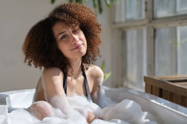 Gülümseyen ve rahatlatan Afro-Amerikan kadın köpük dolu bir küvette banyo yapıyor. Harika bir zaman. Yaşam tarzı insan kavramı