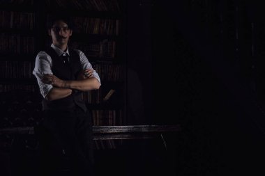 Karanlık bir kütüphane odasında düşünceli bir adam gece ayakta duruyor. Bıyıklı şık bir adam bileziği düzeltiyor..