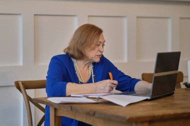 Üst düzey kadın mutfağında web sörfü yapmak için dizüstü bilgisayar kullanıyor. Kıdemli iş ve sosyal güvenlik kavramı. Olgun bir kadın iş yerinde oturmuş, bir ofiste defter bilgisayarı yazıyor..