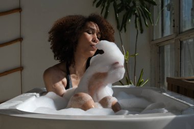 Gülümseyen ve rahatlatan Afro-Amerikan kadın köpük dolu bir küvette banyo yapıyor. Harika bir zaman. Yaşam tarzı insan kavramı