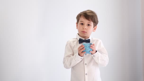Asijský malý chlapec v bílé košili s motýlkem třese prázdnou prasečí bankou, bez peněz koncepce, infantilní postoj k financování — Stock video
