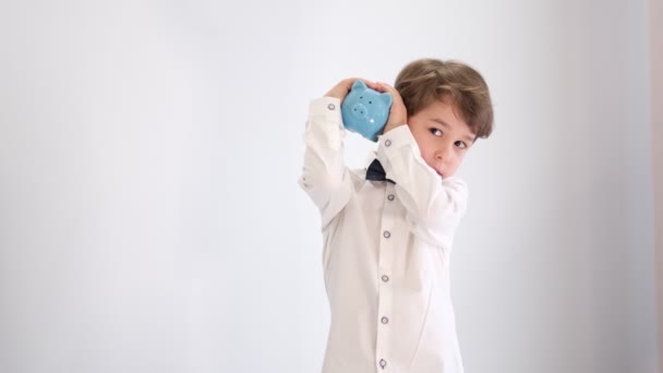 Asiático pequeño niño en una camisa blanca con una pajarita sacude una hucha vacía, sin concepto de dinero, actitud infantil para financiar — Vídeos de Stock