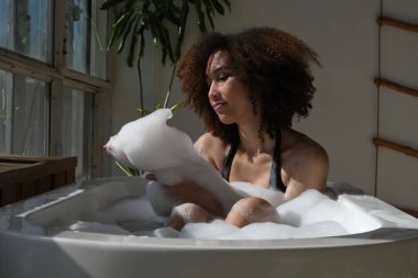 Gülümseyen ve rahatlatan Afro-Amerikan kadın köpük dolu bir küvette banyo yapıyor. Harika bir zaman. Yaşam tarzı insan kavramı