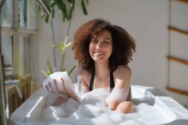 Gülümseyen ve rahatlatan Afro-Amerikan kadın köpük dolu bir küvette banyo yapıyor. Harika bir zaman. Yaşam tarzı insan kavramı