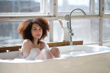 Gülümseyen ve rahatlatan Afro-Amerikan kadın köpük dolu bir küvette banyo yapıyor. Harika bir zaman. Yaşam tarzı insan kavramı