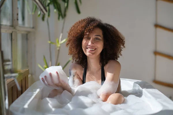 Χαμογελώντας και χαλαρώνοντας Αφροαμερικανή γυναίκα που κάνει μπάνιο σε μια μπανιέρα γεμάτη αφρό. Καταπληκτικές στιγμές. lifestyle άνθρωποι έννοια — Φωτογραφία Αρχείου