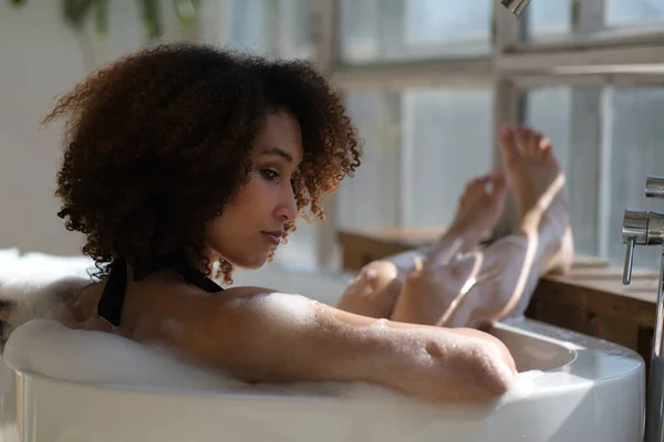 Vista trasera chica negra rizado por la que se extiende, relajarse en el baño en el interior. mujer joven que se relaja en bañera caliente con espuma y burbujas. Mujer cansada disfrutando del descanso mimarse. — Foto de Stock