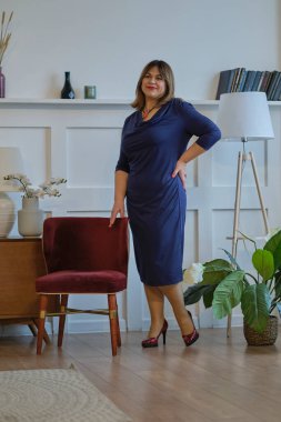 Evde abartılı giyinen bir bayan var. Kendine güvenen olgun bir kadının portresi. Gülümseyen kadın kameraya bakıyor. Başarılı orta yaşlı kadın gülümseyerek evde.