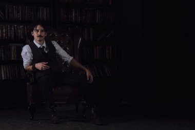 Karanlık bir kütüphane odasında düşünceli bir adam gece ayakta duruyor. Bıyıklı şık bir adam bileziği düzeltiyor..