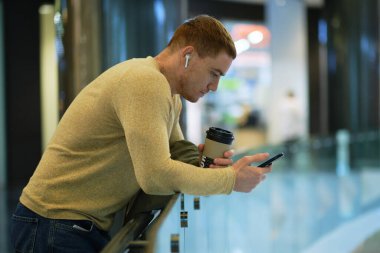 Genç adam akıllı telefon kullanıyor ve bluetooth kulaklıkla müzik dinliyor. Sonbahar ceketli yalnız adam kahve içer..