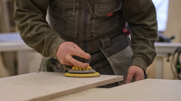 De cerca. Carpintero en taller pule tablero de madera con una lijadora orbital eléctrica. Concepto de carpintería y mobiliario. — Foto de Stock