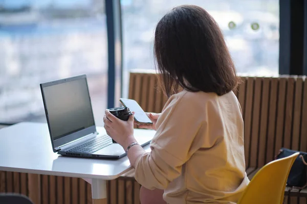 Egy nő telefonál és nézi a képernyőjét. Fiatal hipszter nő ül a kávéban, kávét iszik és laptopot használ.Az asztalon laptop. Lány böngészés internet, chat, blogolás. — Stock Fotó