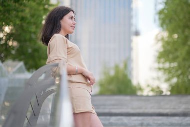 Kadın merdivenlerde duruyor ve yan tarafa bakıyor, arka planda gökdelenler ve ağaçlar.