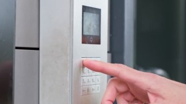 Erkek, bina girişinde dahili telefon kullanıyor. Elektronik kod kilidini açar. Man Hand güvenlik sistemi kodunu giriyor, işaret parmağı dahili iletişim cihazı giriş kapısı olan düğmeye basıyor.