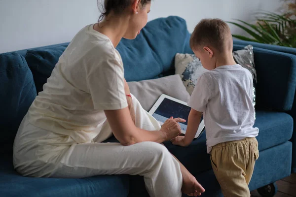Anya és fia tablettel otthon. Anya mutatja média tartalom on-line a fia egy tabletta ül egy kanapén a nappaliban egy házban belső — Stock Fotó