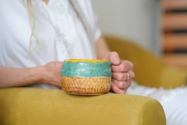 Femme méconnaissable en vêtements blancs assis sur un canapé jaune et tient une tasse jaune à la main. — Photo