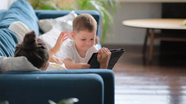 Mãe e filho com tablet em casa. Mãe mostrando conteúdo de mídia on-line para seu filho em um tablet sentado em um sofá na sala de estar em um interior da casa — Fotografia de Stock