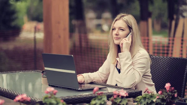 Mulher de negócios sentada à mesa no café, falando no celular enquanto toma notas no caderno. Na mesa laptop e xícara de café. Estudante aprendendo online. Freelancer trabalhando online. — Fotografia de Stock