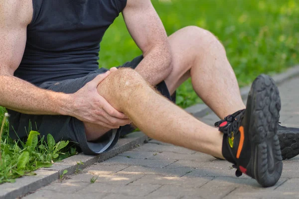 Mężczyzna biegacz dotykający stóp w bólu z powodu zwichniętej kostki. nierozpoznany sportowiec siedzi na ziemi i cierpi na ból kolana — Zdjęcie stockowe