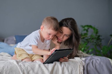 Anne ve oğlu tabletle evde. Anne, evdeki oturma odasındaki bir tabletteki oğluyla internetten medya içeriğini gösteriyor.