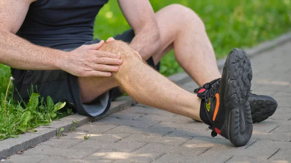 Mężczyzna biegacz dotykający stóp w bólu z powodu zwichniętej kostki. nierozpoznany sportowiec siedzi na ziemi i cierpi na ból kolana — Zdjęcie stockowe