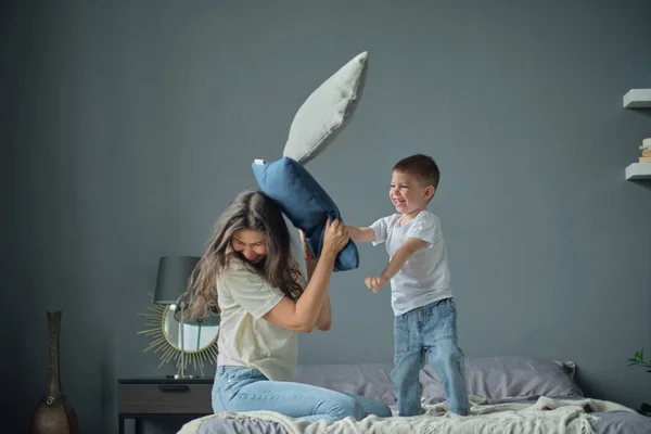 Chico gracioso arroja almohadas a mamá, guerra de almohadas, infancia feliz. Maternidad, concepto de agresión infantil —  Fotos de Stock