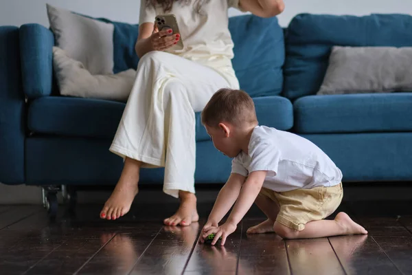 Den söta lilla pojken leker på golvet med en bil, medan mamma eller barnflicka surfa i din smartphone i soffan — Stockfoto