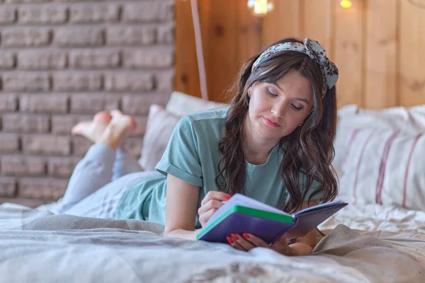 Lifestyle weiches Bild einer hübschen jungen Frau, die auf ihrem gemütlichen Bett sitzt und Notizen in ihr Tagebuch macht. Herbststimmung. — Stockfoto