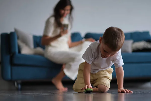 Bild av ung söt liten pojke som leker på golvet med bil leksaker och rita medan hans mamma sitter på en soffa. — Stockfoto
