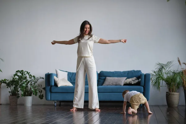 Bonne mère célibataire consciente faisant des exercices matinaux dans la pose de yoga avec l'enfant jouant à la maison. exercices matinaux en pyjama. enfant interfère avec l'entraînement — Photo