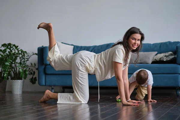 Happy mindful single mother doing morning exercises in yoga pose with kid playing at home. morning exercises in pajamas. child interferes with the workout — Stock Photo, Image