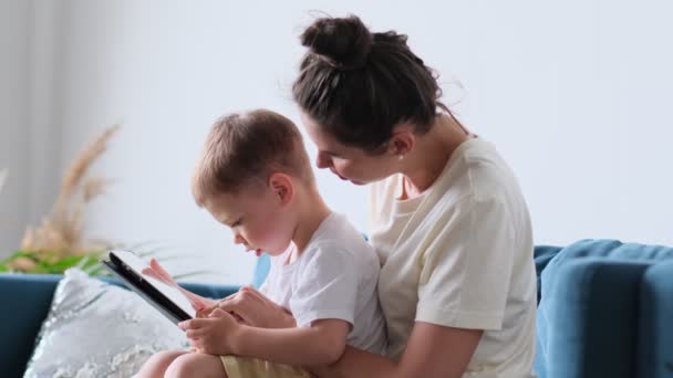 Matka a syn s tabletou doma. Matka zobrazující mediální obsah on-line svému synovi v tabletu sedícím na gauči v obývacím pokoji v interiéru domu — Stock video