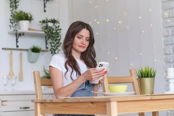 Giovane bella donna usando il cellulare e prendendo un caffè in cucina. — Foto Stock