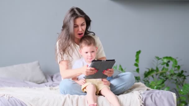 Madre e hijo con la tableta en casa. Madre mostrando contenido multimedia en línea a su hijo en una tableta sentada en un sofá en la sala de estar en el interior de una casa — Vídeo de stock