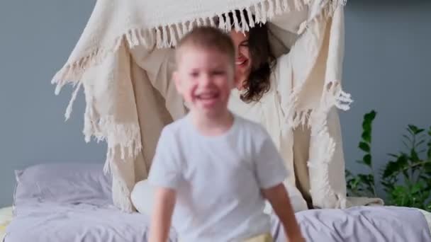 Maman et l'enfant s'amusent dans la chambre sur le lit, jouer sous la couverture. Jeune mère avec son petit fils habillé jouant dans le lit le week-end ensemble, le matin paresseux, scène chaleureuse et confortable. — Video