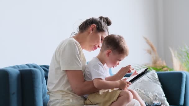 Madre e figlio con tablet a casa. Madre che mostra il contenuto dei media online a suo figlio in un tablet seduto su un divano in soggiorno in un interno della casa — Video Stock