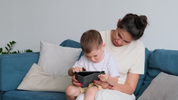 Moeder en zoon met tablet thuis. Moeder tonen media-inhoud on line aan haar zoon in een tablet zitten op een bank in de woonkamer in een huis interieur — Stockvideo