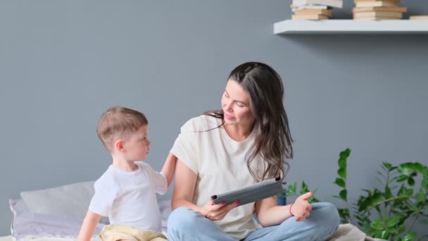 Madre e hijo con la tableta en casa. Madre mostrando contenido multimedia en línea a su hijo en una tableta sentada en un sofá en la sala de estar en el interior de una casa — Vídeo de stock