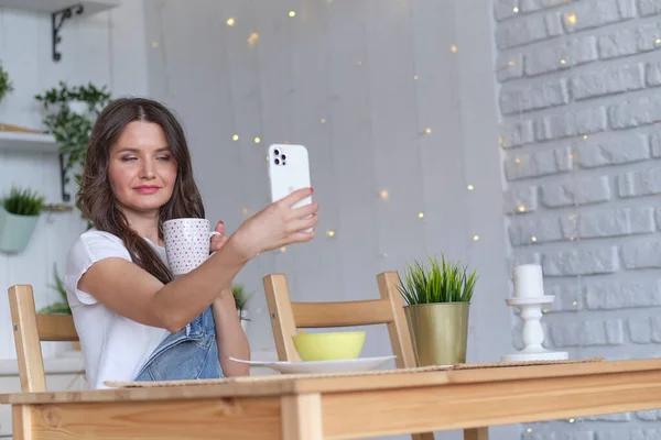 Femminile sorridente utilizzando il telefono cellulare che tiene il caffè a casa in cucina. ordinare cibo attraverso negozi online. acquisto di cibo tramite un'applicazione online — Foto Stock