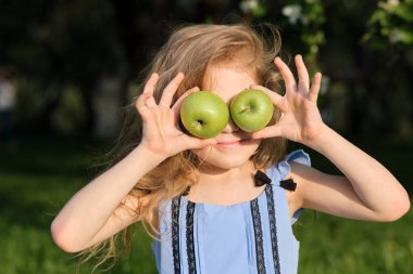 Gülümseyen kız, elmaları gözlerinin önünde, bulanık yeşil arka planda tutuyor.