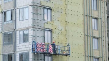 İşçiler bir binanın duvarını yalıtıyorlar. İnşaat alanı. Yabancı işçiler. Yalıtım panelleri. Asılı beşik. Tehlikeli bir iş. Sokakta çalış. Gri wal