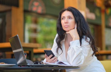 Kahve dükkanındaki olgun kadın. Yazlık terasta oturan kendine güvenen profesyonel bir kadının portresi, iş için dizüstü bilgisayar kullanması, kapalı alanda mutlu bir şekilde gülmesi..