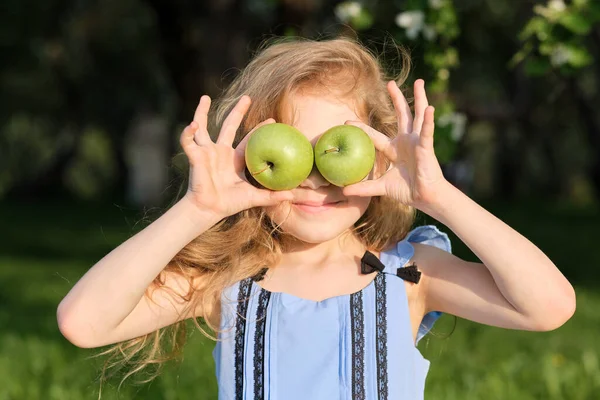 Χαμογελαστό κορίτσι, κρατώντας μήλα μπροστά στα μάτια της πάνω από θολή πράσινο φόντο κήπο — Φωτογραφία Αρχείου
