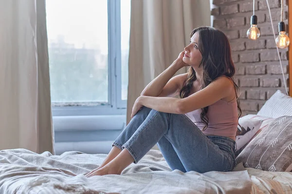 Attraktive Frau mittleren Alters, die sich zu Hause entspannt. schöne Brünette sitzt auf dem Bett und umarmt ihre Knie — Stockfoto