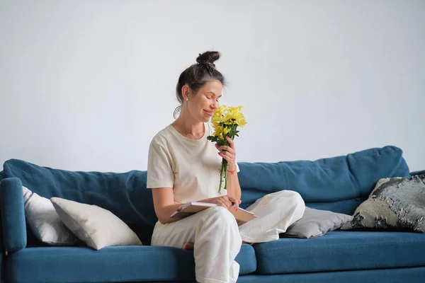 Mutlu kadın evinde, kanepede otururken çiçeklerin kokusunu içine çekiyor.. — Stok fotoğraf