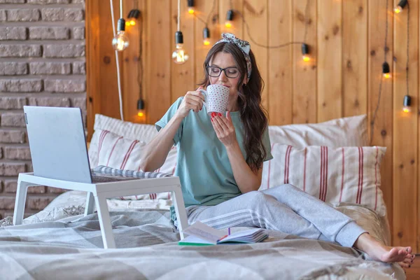 Urocza kobieta w piżamie siedzi na łóżku i pije kawę, przed laptopem, na którym pracuje lub dokonuje zakupów online. koncepcja freelancingu lub pracy zdalnej — Zdjęcie stockowe